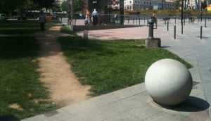 A stone walkway into a larger plaza is partially obstructed by a large stone sphere; walkers have worn their own path around it in the nearby grass.